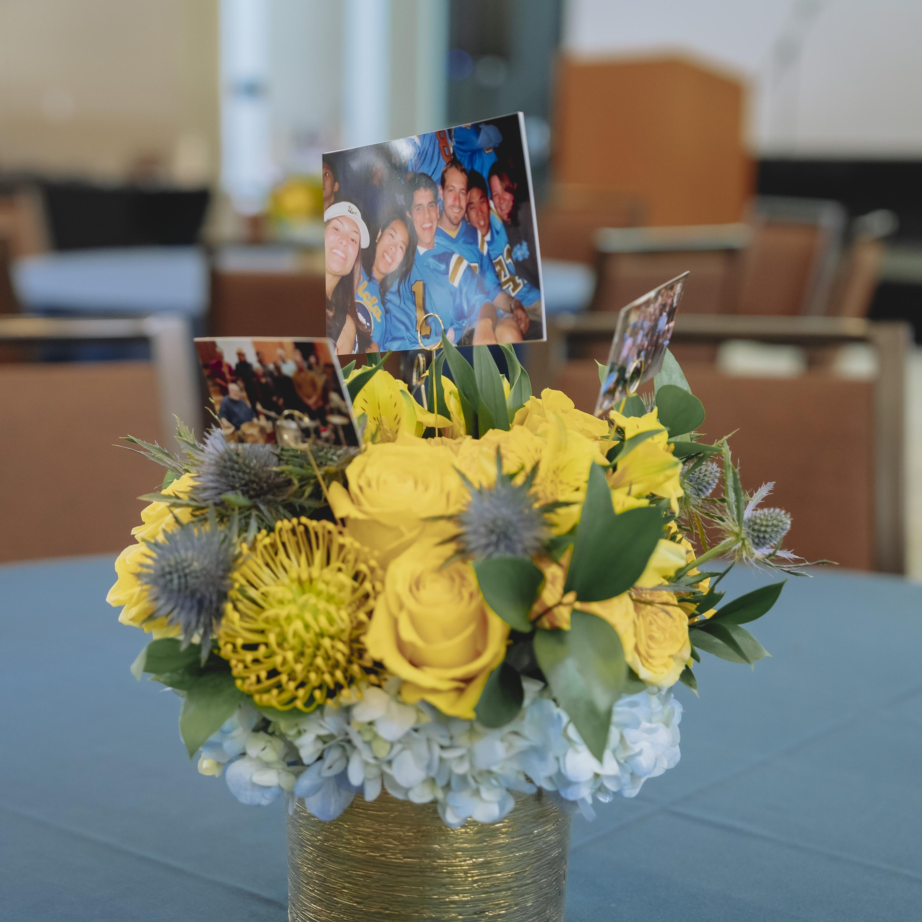 A golden ribbon wrapped vase centerpiece with yellow roses and photos of ucla staff on sticks