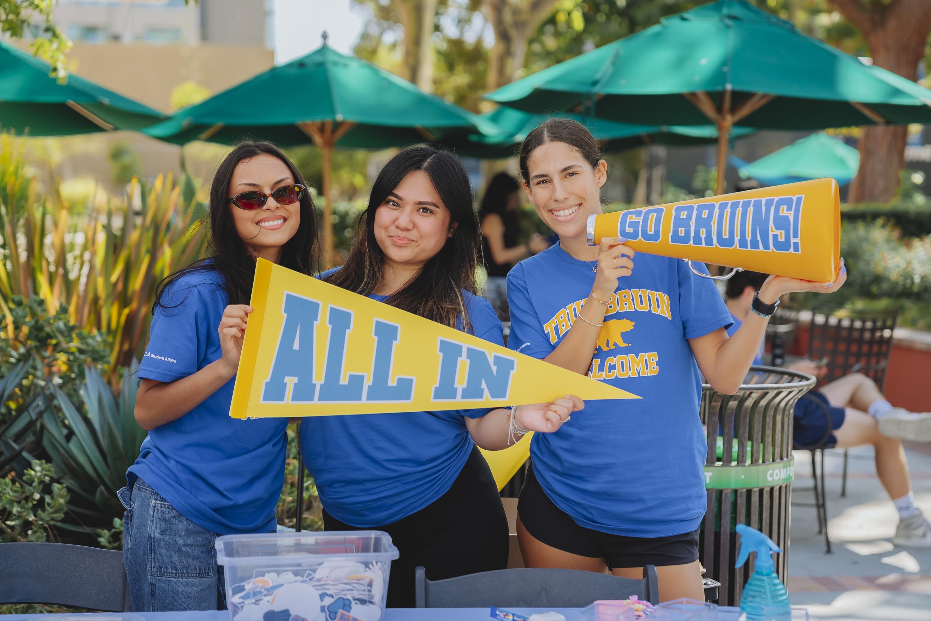 True Bruin Ambassadors holding a yellow pennant that says "ALL IN" and a megaphone that says "Go Bruins!"